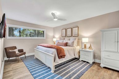 Sunlit coastal-style master bedroom with white wooden bed, layered neutral bedding and rust throw, matching white nightstands and armoire, blue striped rug, ceiling fan and armchair by a window overlooking trees.