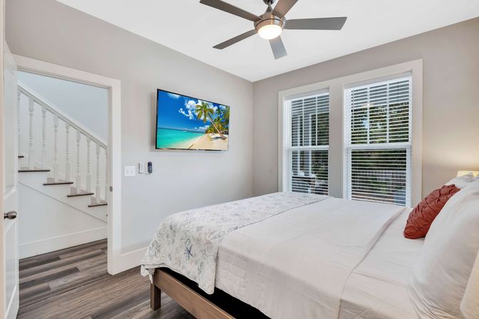 Bright modern coastal-style bedroom with queen bed and white linens, ceiling fan, wall-mounted TV showing a tropical beach, wood floors and double windows with blinds