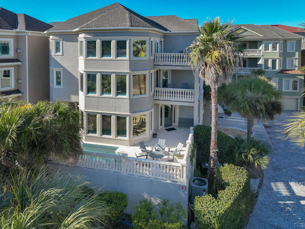Three-story coastal vacation home with wraparound windows, balconies, small plunge pool and patio seating, palm trees and neighboring beach houses under a bright blue sky