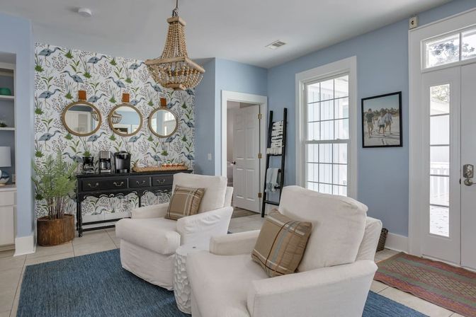 Bright coastal cottage living room with light blue walls, botanical bird wallpaper framed by three round mirrors, a beaded chandelier above a black console with a coffee station, two white armchairs with plaid pillows on a blue rug, and large windows letting in natural light.