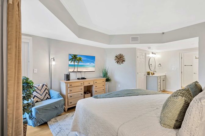 Sunny beach-inspired master bedroom with tray ceiling, king bed and textured pillows, light wood dresser with wall TV showing a tropical beach, blue armchair, potted plants and adjoining en-suite bathroom.