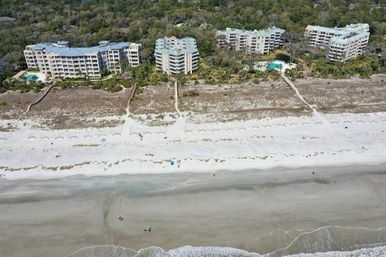Aerial view of beachfront condominium complex with pools and wooden walkways over dunes leading to a wide white-sand beach and gentle ocean waves, scattered beachgoers on the shore.