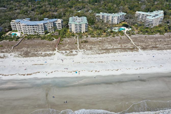 Aerial view of beachfront condominium complex with pools and wooden walkways over dunes leading to a wide white-sand beach and gentle ocean waves, scattered beachgoers on the shore.