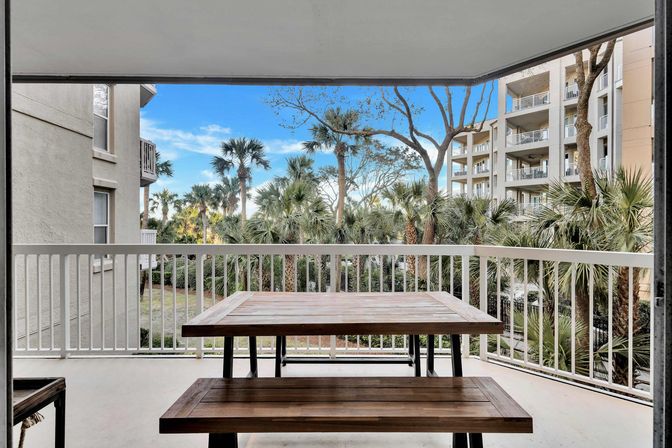 Covered coastal condo balcony with wooden picnic table and bench, white railing overlooking palm trees and neighboring multi-story buildings under a blue sky