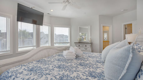 Sunlit coastal-style master bedroom with blue floral bedding, pale blue pillows, neatly folded white towels on the bed, three large windows, wall-mounted flat-screen TV, dresser with mirror and visible en-suite bathroom
