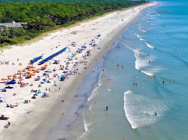 Aerial view of a long sandy beach with colorful umbrellas and sunbathers, people wading in clear blue ocean waves along a green tree-lined coastline — lively coastal vacation scene.