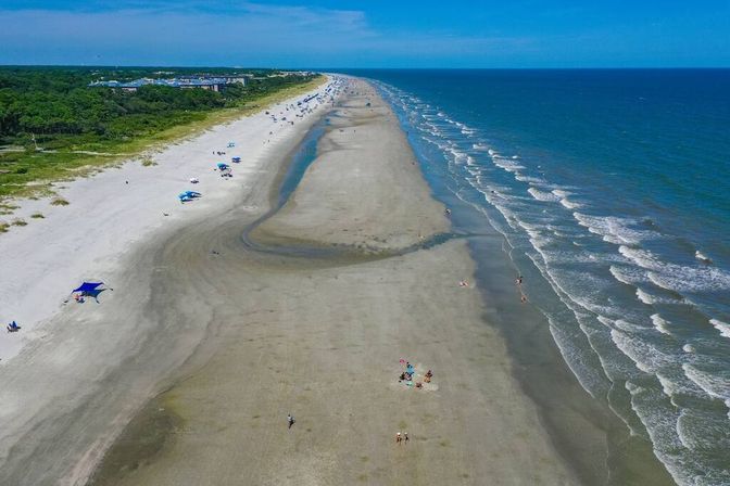 Aerial view of a sunny, wide sandy beach with a curving tidal channel, scattered umbrellas and beachgoers, green dunes to the left and blue ocean waves breaking along the shoreline.