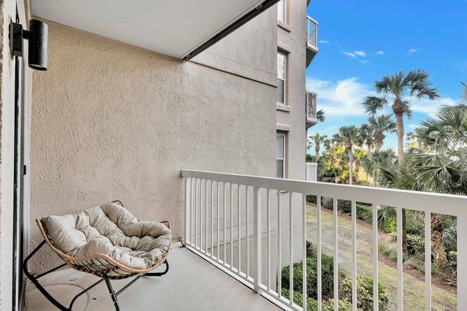 Coastal condo balcony with a cushioned papasan chair, white railing, and tropical palm-tree view under a bright blue sky