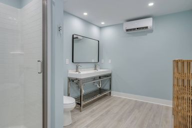 Bright, spa-like light blue bathroom with double sinks on stainless-steel vanity, large mirror, glass walk-in tiled shower, wall-mounted mini-split AC, and wood-look flooring.
