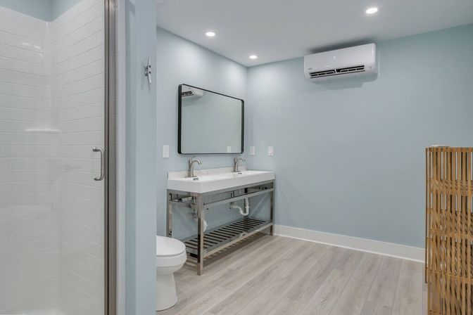 Bright, spa-like light blue bathroom with double sinks on stainless-steel vanity, large mirror, glass walk-in tiled shower, wall-mounted mini-split AC, and wood-look flooring.