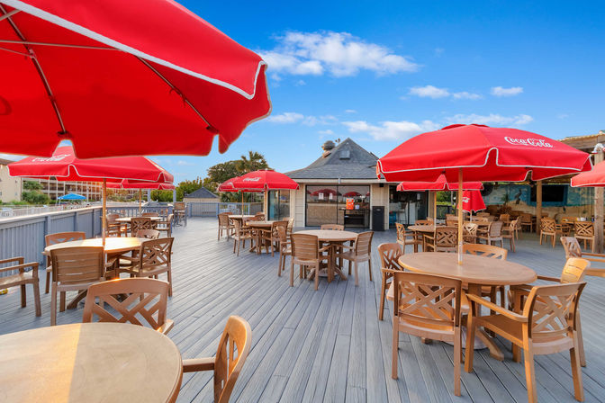Sunny waterfront outdoor dining deck with round wooden tables, chairs, and bright red sun umbrellas on a spacious boardwalk-style patio under a blue sky.