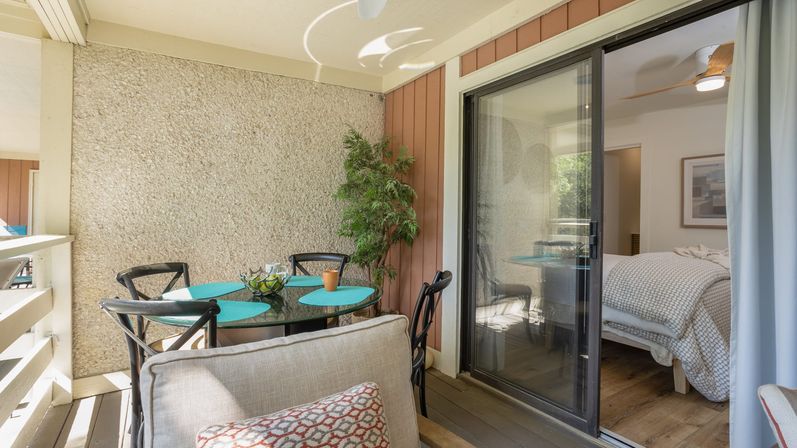 Cozy covered balcony patio with round glass dining table and turquoise placemats, black chairs, potted plant, and sliding glass door into a bedroom.