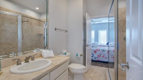 Bright ensuite bathroom with marble countertop vanity and sink, brushed nickel faucet, glass-enclosed tiled shower, white toilet, neutral tile floor, and open door revealing a coastal-themed bedroom with nautical quilt and sunlit windows.