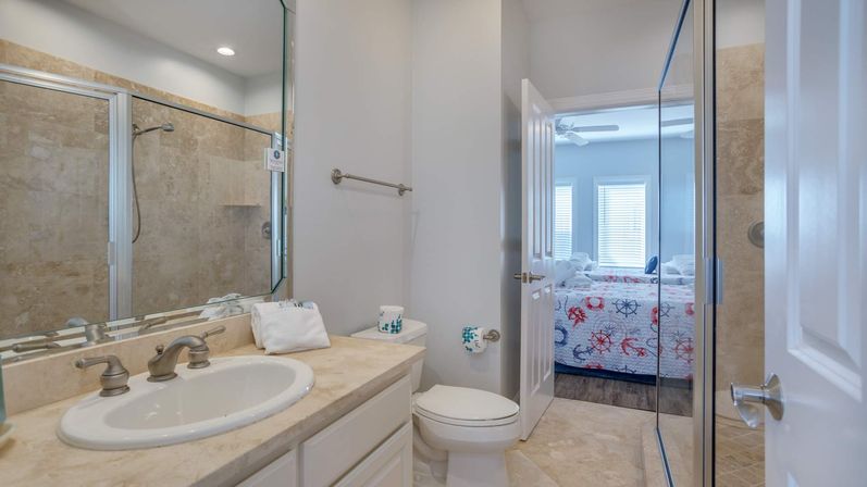 Bright ensuite bathroom with marble countertop vanity and sink, brushed nickel faucet, glass-enclosed tiled shower, white toilet, neutral tile floor, and open door revealing a coastal-themed bedroom with nautical quilt and sunlit windows.
