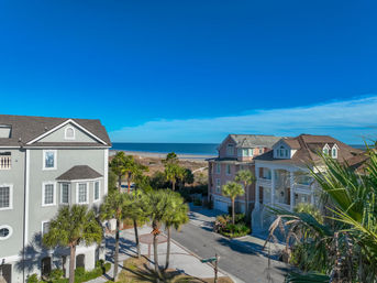 Aerial view of pastel beachfront homes and palm trees along a quiet street opening to a sandy beach and calm ocean under a bright blue sky.