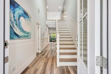 Bright modern coastal-style entry foyer with wood-look plank floors, white wainscoting, a staircase with wood treads, and a large blue wave painting.