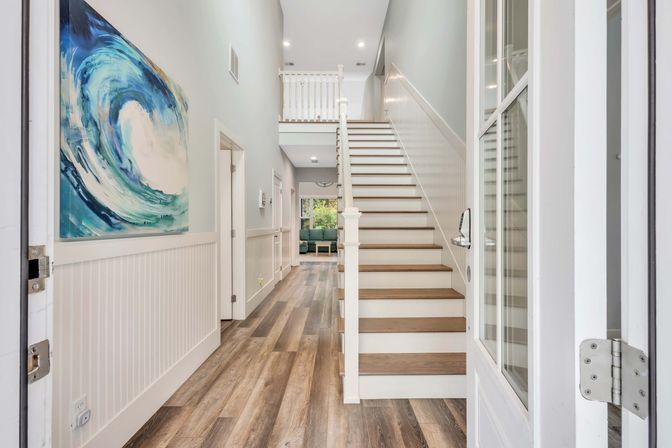 Bright modern coastal-style entry foyer with wood-look plank floors, white wainscoting, a staircase with wood treads, and a large blue wave painting.