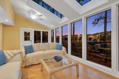 Cozy sunroom with vaulted skylights and floor-to-ceiling windows overlooking pine trees at sunset, cream slipcovered sofas with blue pillows and a wicker glass-top coffee table on warm hardwood floors.
