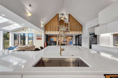 Bright modern coastal open-concept kitchen with white quartz island and brass spring faucet, vaulted ceiling, spiral staircase to loft, and large windows framing palm-tree views.