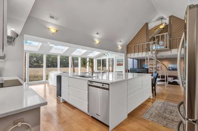 Sunlit open-concept coastal kitchen with large white island, stainless steel appliances, skylights and floor-to-ceiling windows overlooking marshland, hardwood floors and spiral staircase to a loft