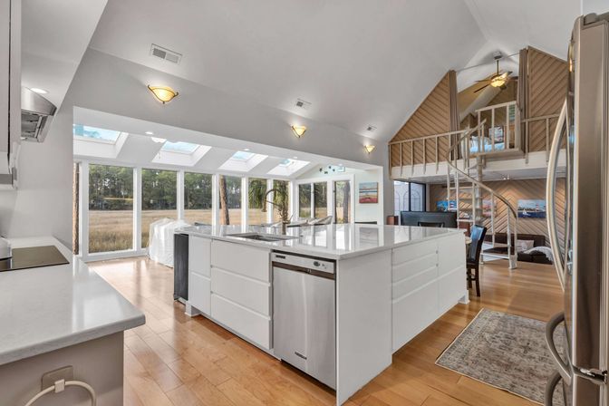 Sunlit open-concept coastal kitchen with large white island, stainless steel appliances, skylights and floor-to-ceiling windows overlooking marshland, hardwood floors and spiral staircase to a loft