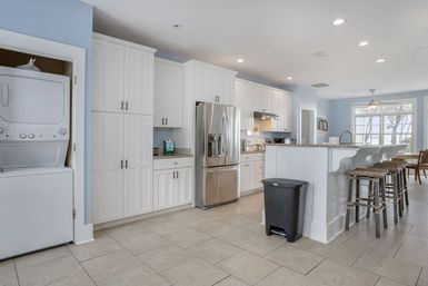 Bright open-concept kitchen with light blue walls, white cabinetry, stainless steel refrigerator, breakfast bar with rustic stools, stacked washer-dryer and large window.