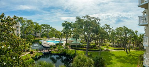 Sunny oceanfront resort grounds with outdoor pool and lounge area beside a pond with fountain, wooden boardwalk, lush live oaks and palm trees, green lawns and condominium balconies overlooking the coastline.