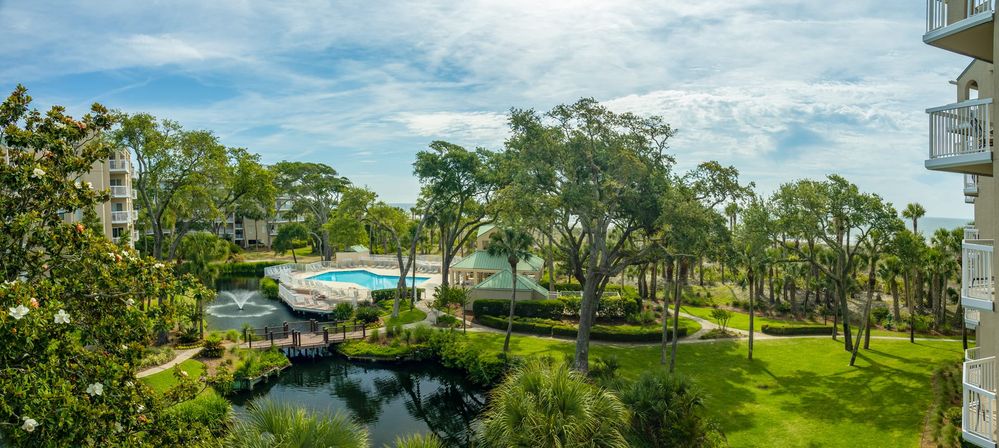 Sunny oceanfront resort grounds with outdoor pool and lounge area beside a pond with fountain, wooden boardwalk, lush live oaks and palm trees, green lawns and condominium balconies overlooking the coastline.