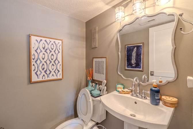 Cozy residential powder room with gray walls, white pedestal sink and toilet with open lid, ornate silver-framed mirror, glass pendant lights, blue framed artwork, teal vases and soap dispenser on the sink and toilet tank.