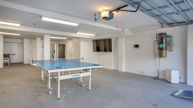Residential garage-style game room with blue ping-pong table on casters, wall-mounted TV, concrete floor, overhead garage door and mounted central vacuum—ready for a match.