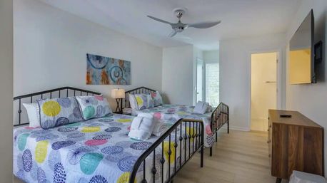 Bright vacation rental bedroom with two colorful patterned double beds on black metal frames, ceiling fan, wall-mounted TV, wooden dresser, en-suite bathroom and shuttered balcony — light wood floors and modern coastal decor.