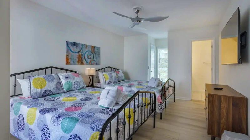 Bright vacation rental bedroom with two colorful patterned double beds on black metal frames, ceiling fan, wall-mounted TV, wooden dresser, en-suite bathroom and shuttered balcony — light wood floors and modern coastal decor.