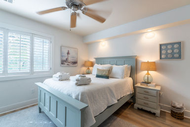 Sunlit coastal cottage bedroom with sea-foam wooden bed, white quilt and rolled towels, lighthouse artwork, glass bedside lamps, plantation shutters and ceiling fan.
