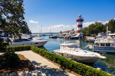 Sunny coastal marina with white yachts and sailboats docked in calm blue water, a manicured waterfront path and green hedges, and a tall red-and-white striped lighthouse under a bright blue sky.