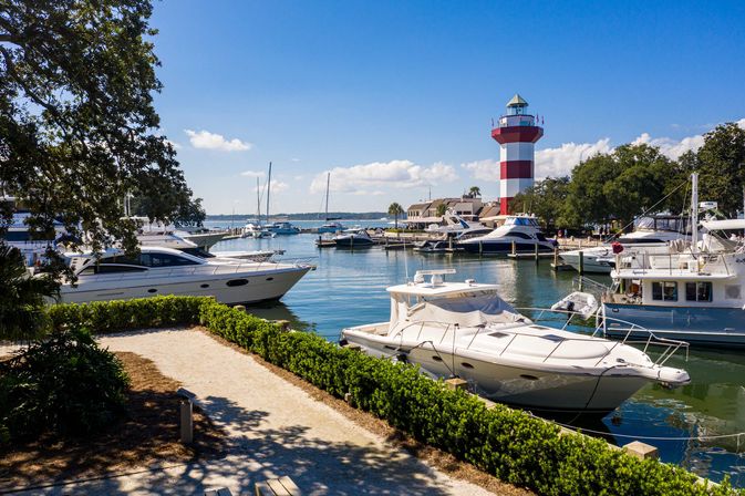 Sunny coastal marina with white yachts and sailboats docked in calm blue water, a manicured waterfront path and green hedges, and a tall red-and-white striped lighthouse under a bright blue sky.