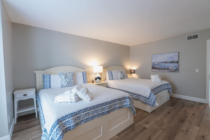 Airy coastal-style guest bedroom with two white beds in blue-and-white quilts, bedside lamps, folded towels, seascape artwork and wood flooring.