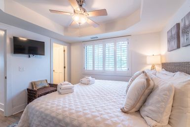 Sunny coastal-style bedroom with a large bed in white quilted bedding and decorative pillows, ceiling fan, wall-mounted TV, wicker chair with a decorative cushion, plantation-shutter window, and bedside lamp.