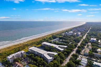 Aerial view of a sunny sandy coastline and blue ocean, with beachfront condos and resorts behind dune vegetation and a tree-lined coastal road stretching into the distance.