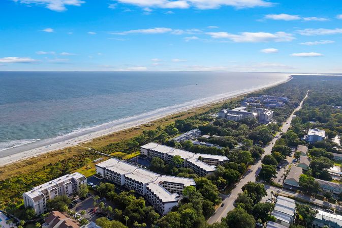 Aerial view of a sunny sandy coastline and blue ocean, with beachfront condos and resorts behind dune vegetation and a tree-lined coastal road stretching into the distance.