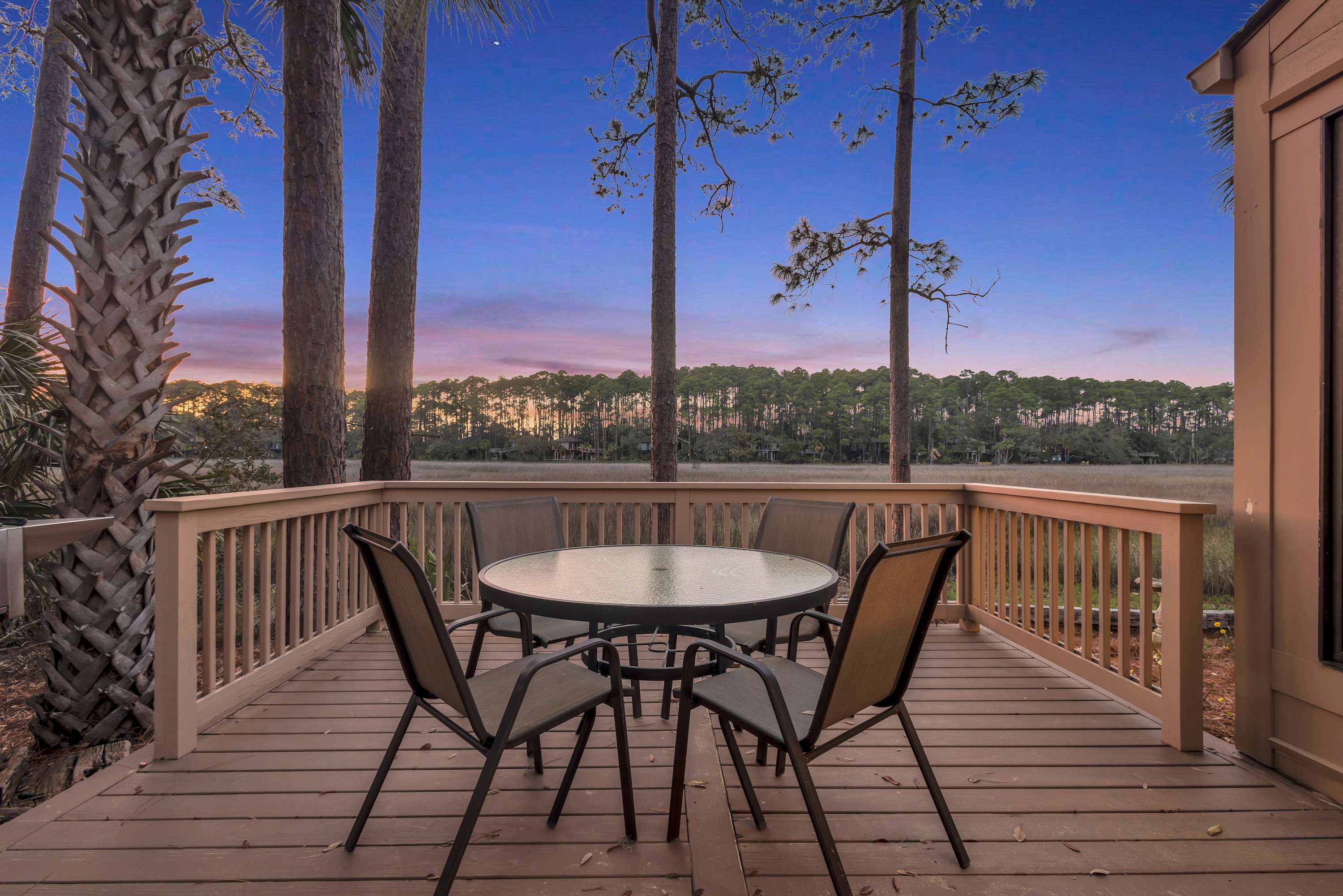 Wooden waterfront deck with a round glass patio table and four chairs overlooking a marsh and pine tree line at sunset under a purple-pink sky.