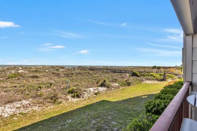 Sunny oceanfront balcony overlooking sandy coastal dunes, green shrubs and a wooden boardwalk leading to the beach under a bright blue sky.