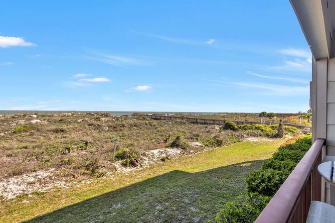 Sunny oceanfront balcony overlooking sandy coastal dunes, green shrubs and a wooden boardwalk leading to the beach under a bright blue sky.
