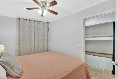 Cozy small bedroom interior with peach quilted bedspread, gray accent pillows, geometric-pattern curtains, wooden-blade ceiling fan and open built-in shelving closet on light hardwood floors.