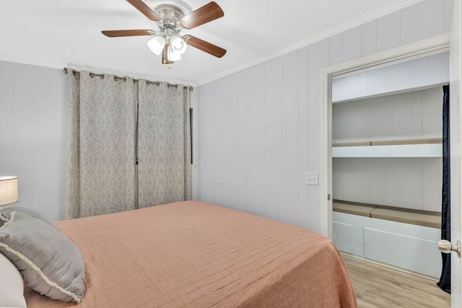 Cozy small bedroom interior with peach quilted bedspread, gray accent pillows, geometric-pattern curtains, wooden-blade ceiling fan and open built-in shelving closet on light hardwood floors.