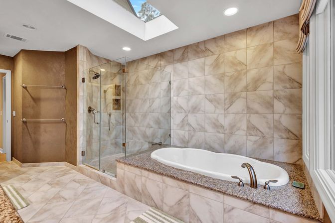 Spa-like master bathroom with marble-look tile walls and floor, glass-enclosed shower, granite-topped soaking tub with bronze fixtures, skylight and towel racks.
