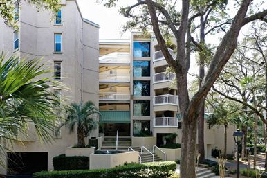 Beige multi-story condominium with stacked balconies and exterior stairways, framed by palm and oak trees around a landscaped courtyard with a small fountain