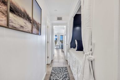 Coastal-style hallway with tile floor, beach-dune wall art, built-in curtained window seat with navy pillows, and a balcony opening to an ocean view.