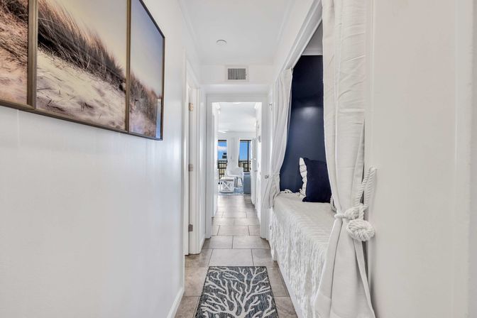 Coastal-style hallway with tile floor, beach-dune wall art, built-in curtained window seat with navy pillows, and a balcony opening to an ocean view.