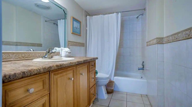 Cozy residential bathroom with oak vanity and marble countertop, sink and mirror, tiled floor, white bathtub with shower curtain and toilet