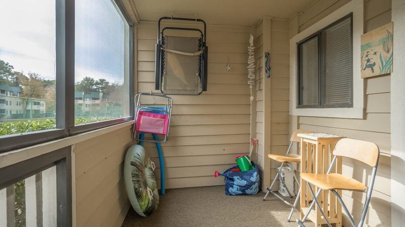 Screened balcony with stacked folding beach chairs, inflatable tube and sand toys in a tote, plus two wooden stools—cozy coastal condo porch